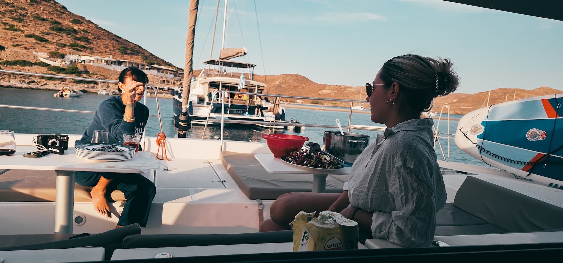 Two sailors at the cockpit table of a catamaran at anchor in a Greek bay