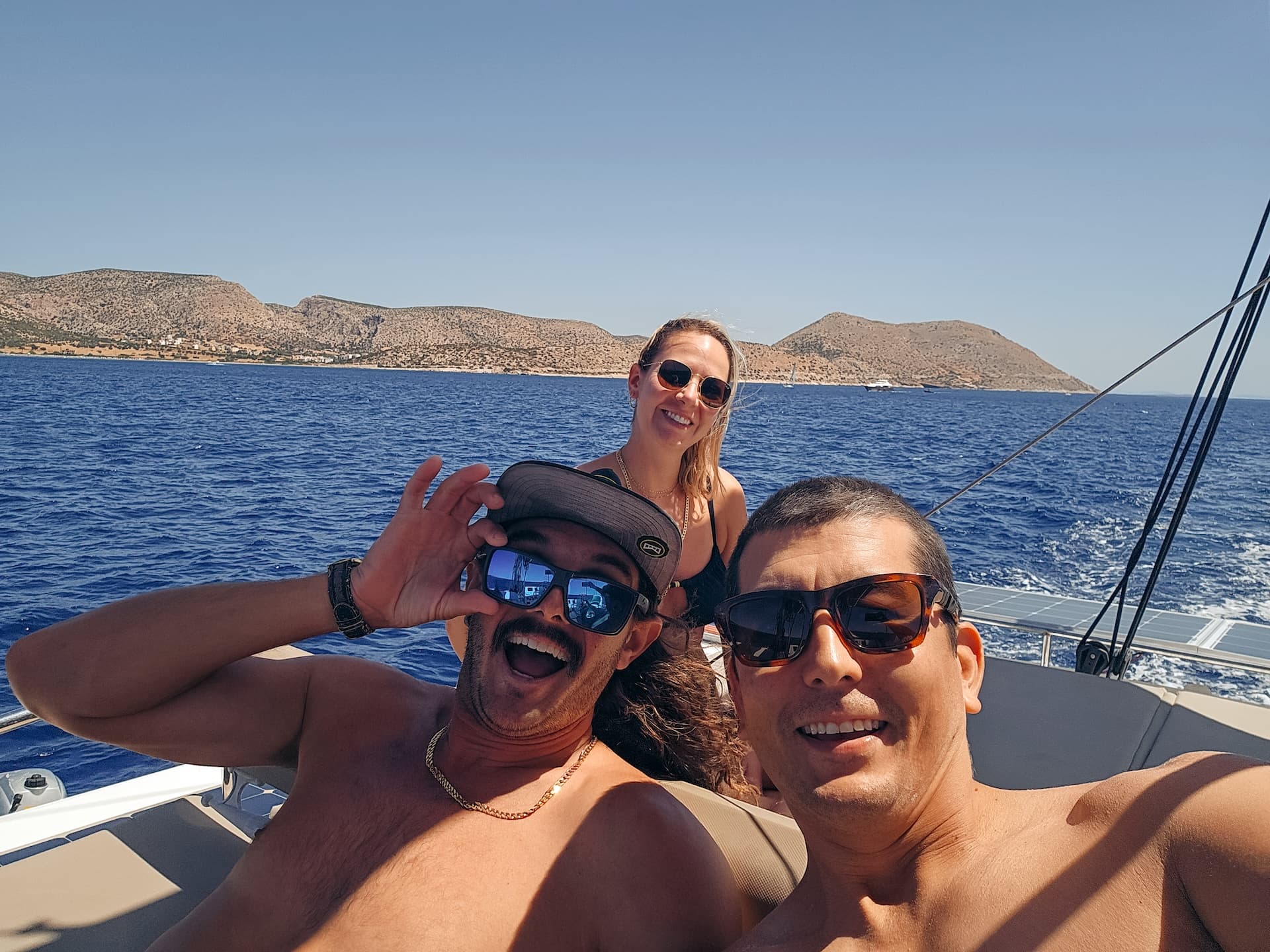 Three Sailing Virgins sailors grinning at the camera on deck, Greek islands behind them